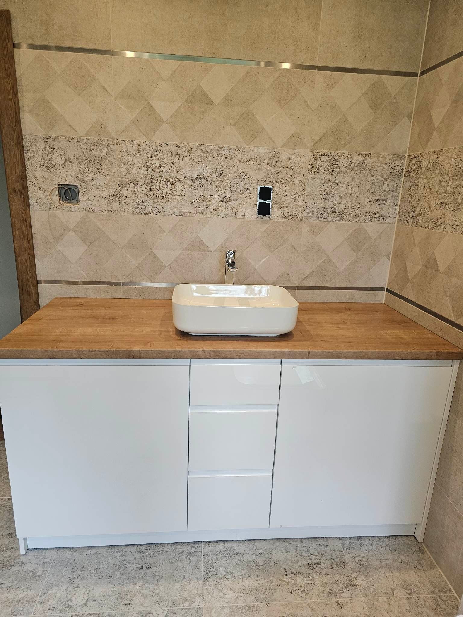 Nowoczesna łazienka z białą szafką pod umywalkę z trzema szufladami, drewniany blat, prostokątna umywalka nablatowa, neutralne płytki na ścianach i podłodze, widoczne gniazdka elektryczne.