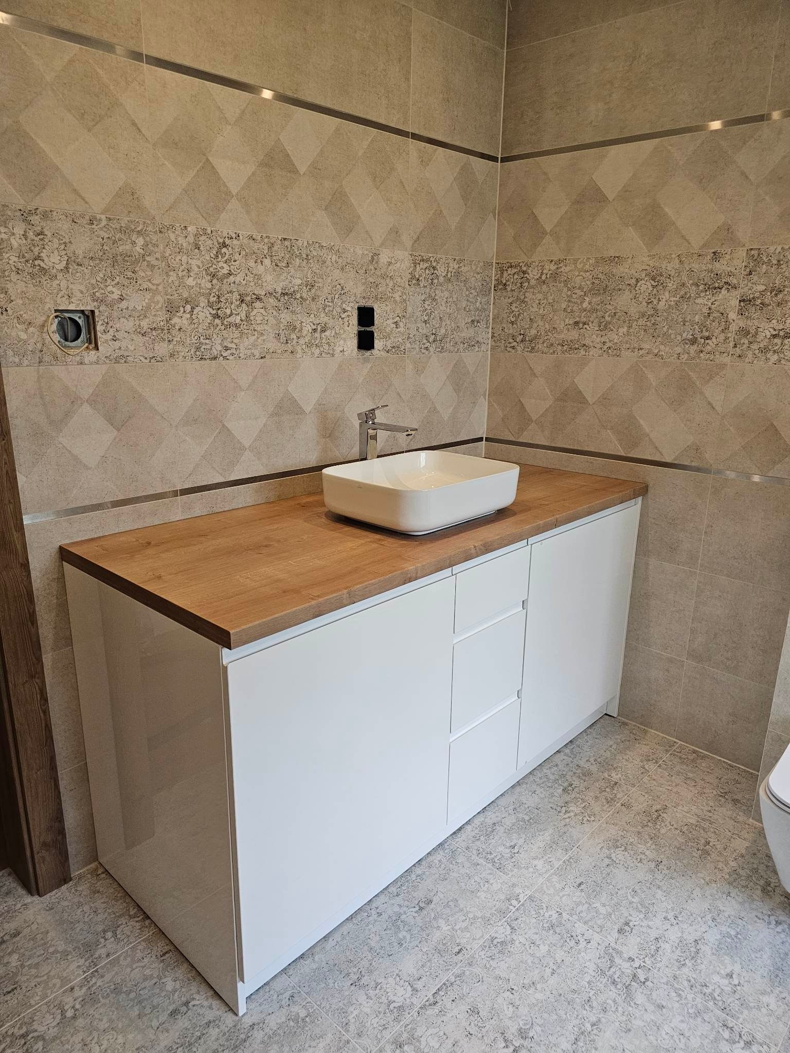 Łazienka z białą szafką umywalkową z drewnianym blatem, kwadratowa umywalka nablatowa i beżowe płytki na ścianach.