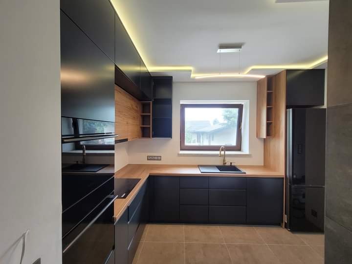 Nowoczesna kuchnia z czarnymi frontami szafek, drewnianymi blatami i akcentami z naturalnego drewna, oświetleniem LED pod sufitem i minimalistyczną lampą wiszącą nad zlewem.