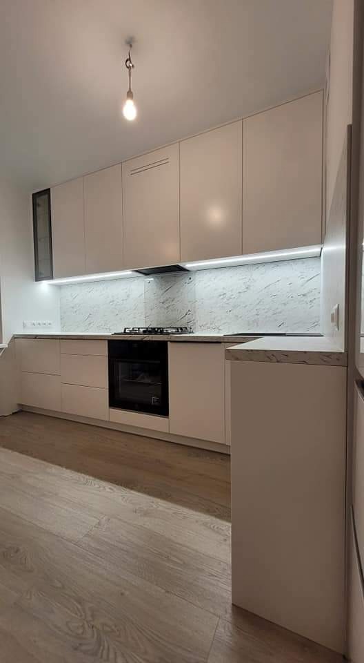 Nowoczesna, jasna kuchnia z białymi frontami szafek, blatem imitującym marmur i subtelnym oświetleniem LED pod górnymi szafkami, z widoczną pojedynczą żarówką na suficie.