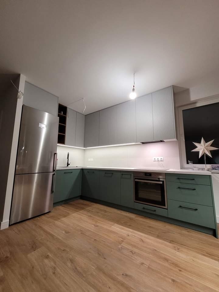 Nowoczesna kuchnia w odcieniach szarości i zieleni z drewnianą podłogą, oświetleniem LED pod szafkami górnymi i wiszącą żarówką bez klosza.