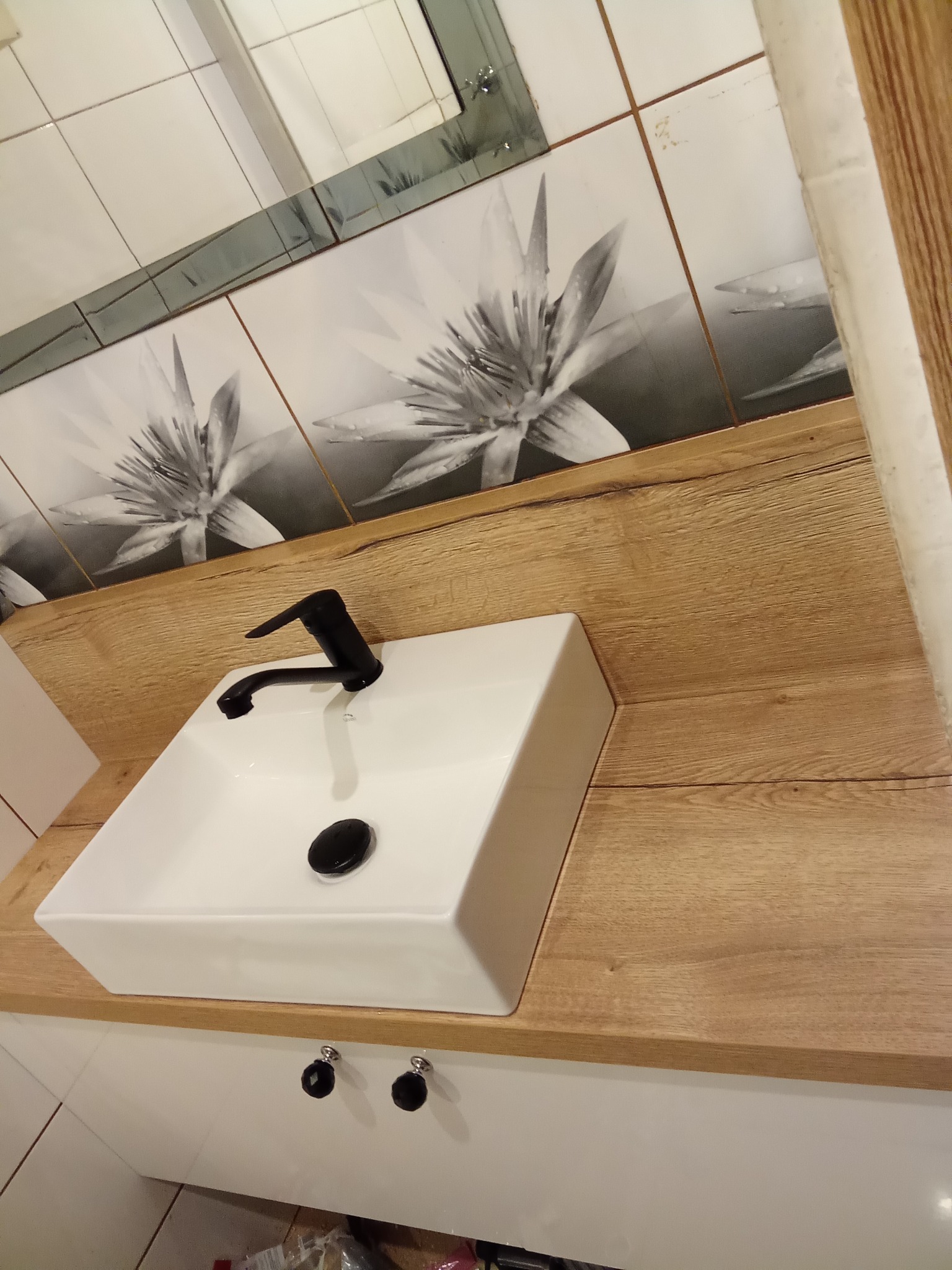 Nowoczesna łazienka: biała, prostokątna umywalka z czarną armaturą na blacie z jasnego drewna. W tle płytki z motywem kwiatowym i lustro.