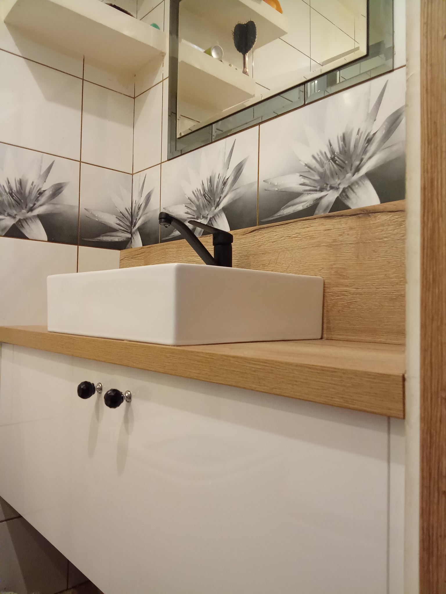 Biała, prostokątna umywalka nablatowa z czarną armaturą na blacie z jasnego drewna. Pod spodem biała szafka z czarnymi gałkami. W tle płytki z motywem kwiatowym.