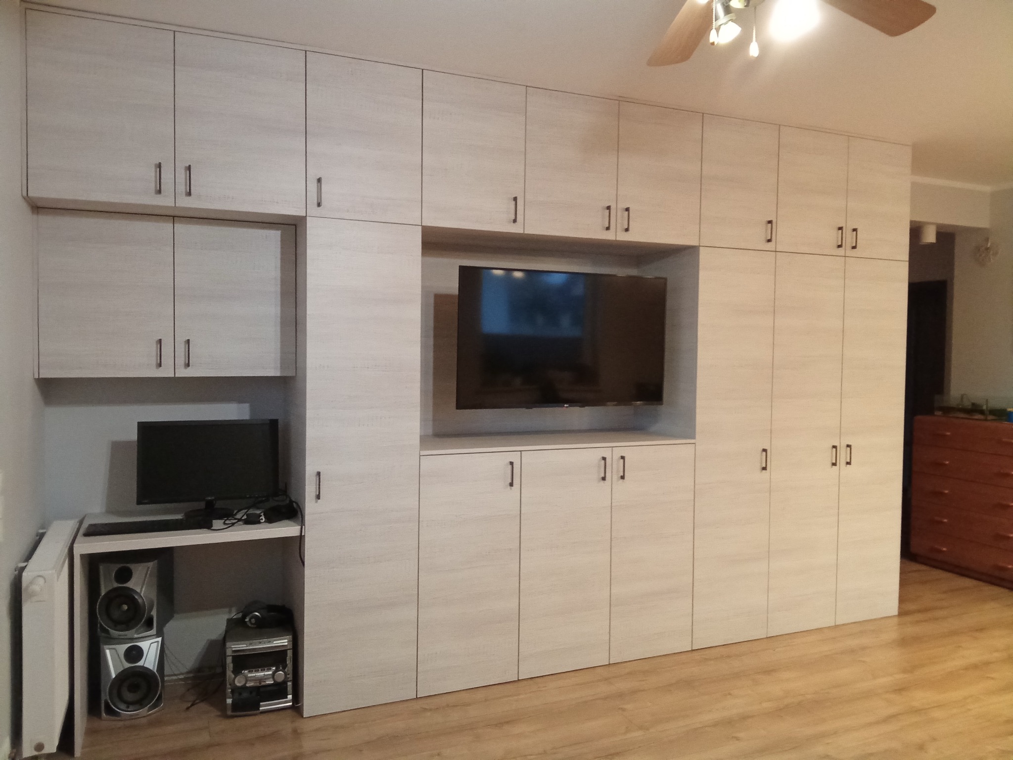 Jasna zabudowa meblowa z szafkami i miejscem na TV oraz biurko z komputerem. Minimalistyczne uchwyty, drewnopodobny dekor. Funkcjonalne rozwiązanie do salonu.