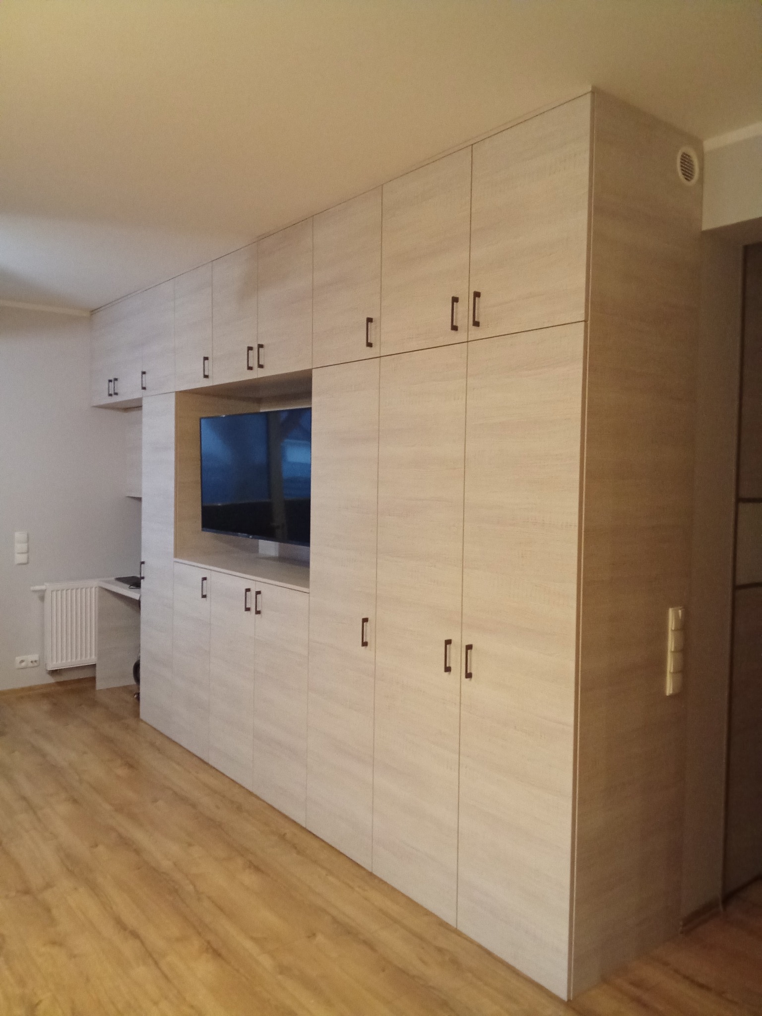 Jasna zabudowa meblowa z szafkami i miejscem na telewizor, dopasowana do wnętrza. Minimalistyczne uchwyty, drewnopodobny dekor. Widok na podłogę i fragment biurka.