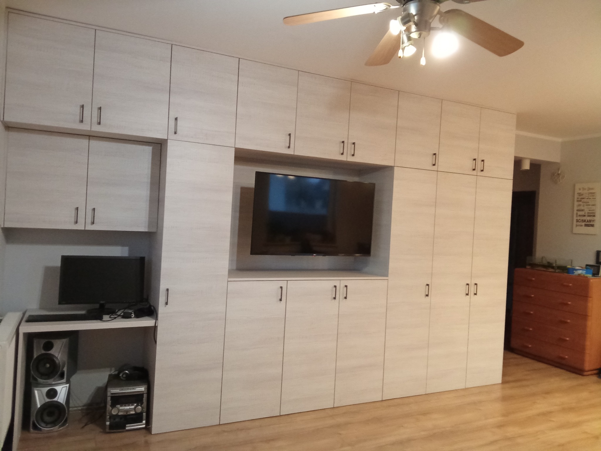 Jasna zabudowa meblowa z szafkami i wnęką na telewizor. Po lewej stronie biurko z komputerem i sprzętem audio. Widoczny parkiet i sufit z wentylatorem.