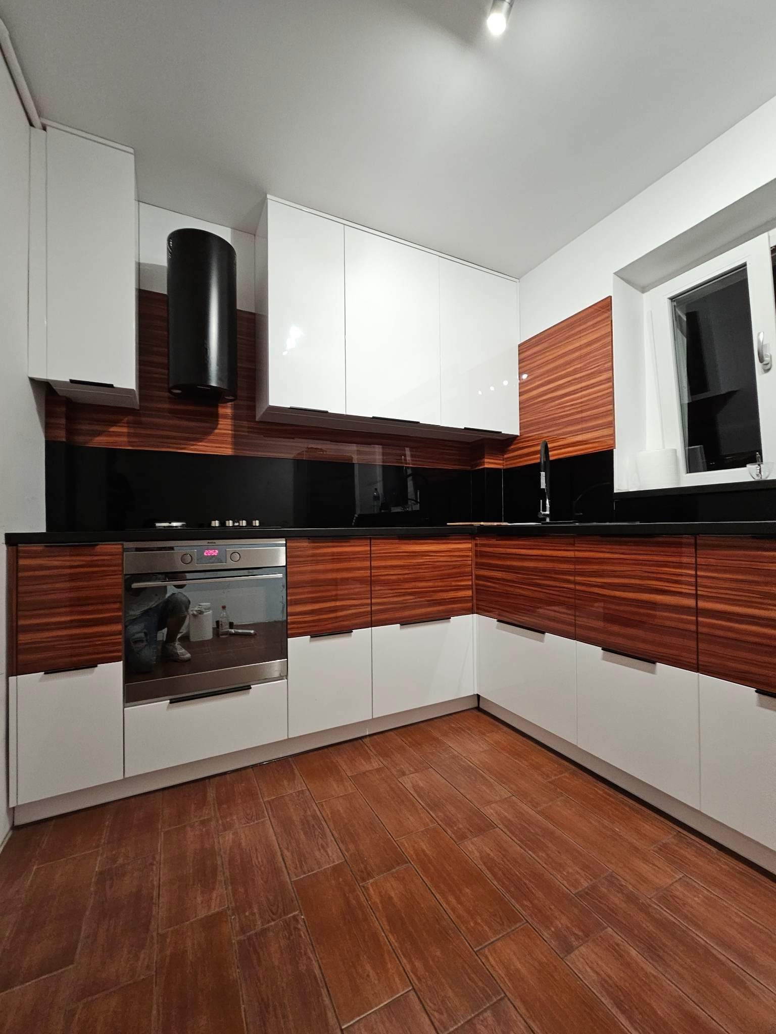 Nowoczesna kuchnia w Nowym Tomyślu: biało-brązowe szafki z fornirem, czarny blat i szkło, drewnopodobna podłoga. Minimalistyczny design z piekarnikiem w zabudowie.