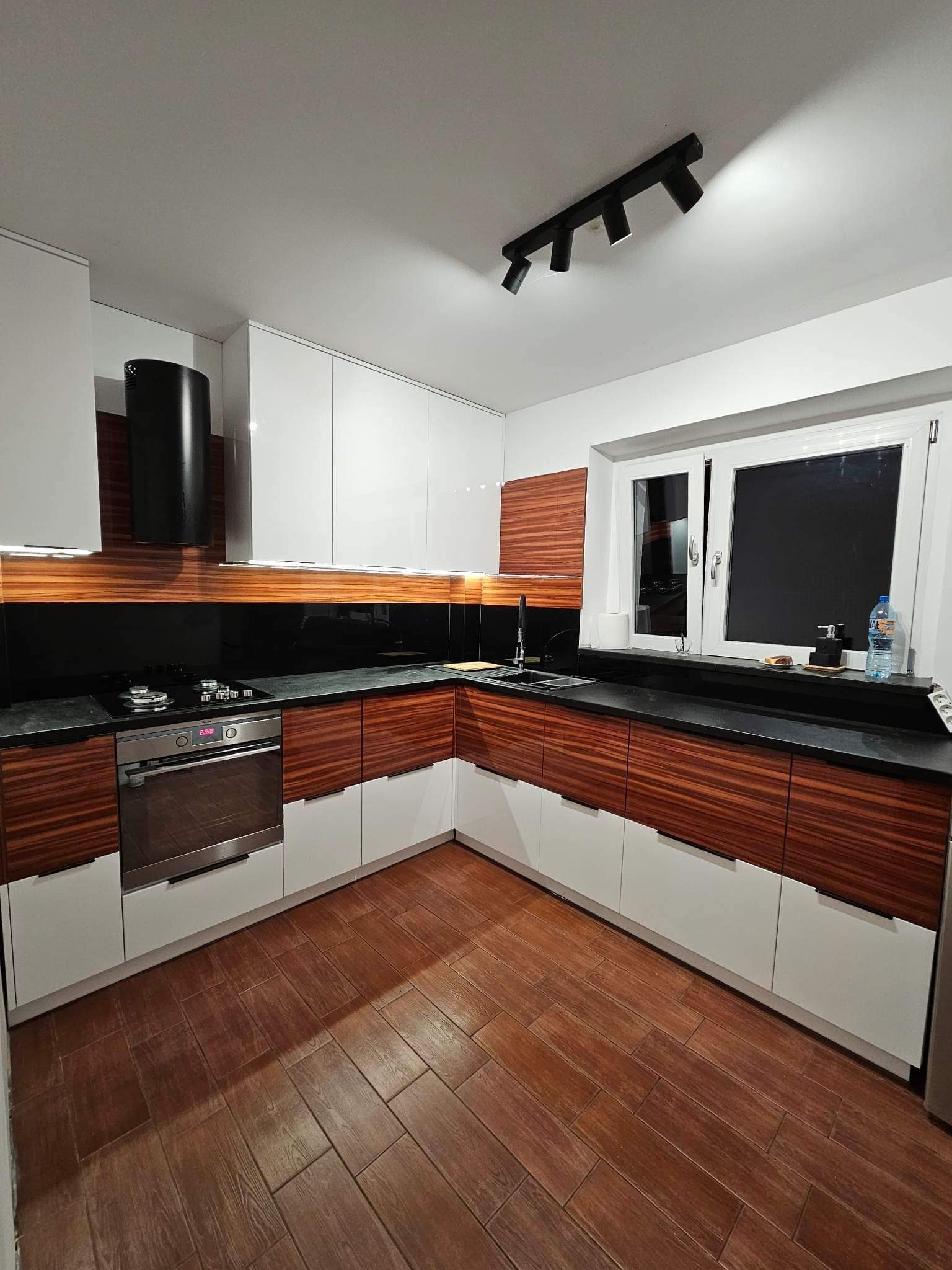 Nowoczesna kuchnia z białymi, lakierowanymi szafkami górnymi i dolnymi z fornirem w kolorze drewna. Czarny blat i panel nad blatem. Oświetlenie punktowe.