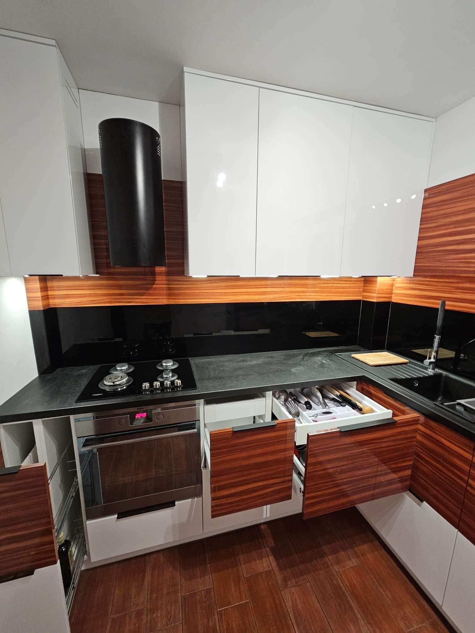 Nowoczesna kuchnia z białymi szafkami i drewnopodobnymi frontami. Otwarta szuflada z organizerem na sztućce. Czarny blat i panel nad kuchenką. Minimalistyczny okap.