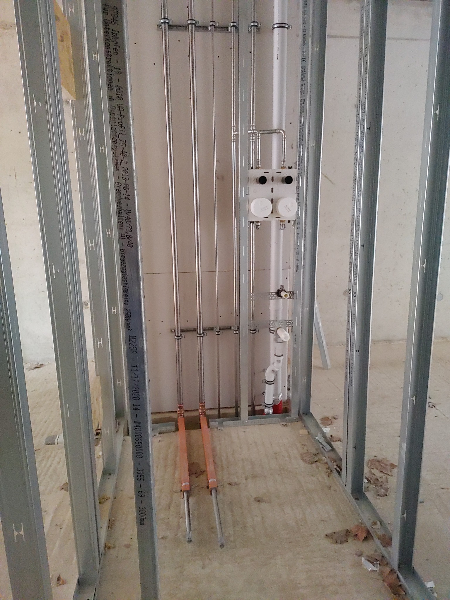 Wewnętrzne ramy ścian działowych z metalowymi profilami, pionowe rury instalacji wodnej i grzewczej, biała zabudowa podtynkowa z zaworami, widoczne elementy instalacji podłogowej.