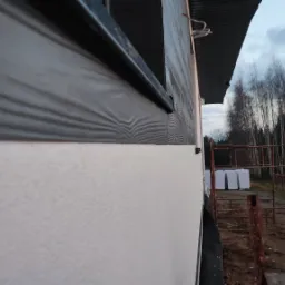 Fragment elewacji budynku z szarą okładziną imitującą drewno i białą, chropowatą ścianą, widok z boku, w tle drzewa bez liści i dostawczy samochód.