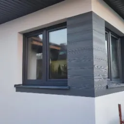 Wykończona elewacja domu z szarymi panelami imitującymi drewno wokół okien i białym tynkiem, widoczne okna z szarymi ramami, na pierwszym planie rusztowanie.