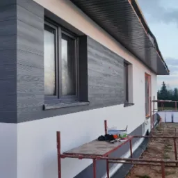 Elewacja domu jednorodzinnego z szarą okładziną imitującą drewno, widoczne rusztowanie i puszki z farbą, drzewa w tle.