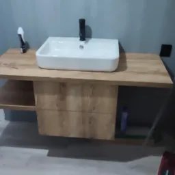 Nowoczesna, prostokątna umywalka nablatowa z czarną armaturą, zamontowana na drewnianej szafce z półką w łazience z szarą ścianą, widoczne narzędzia przy ścianie.