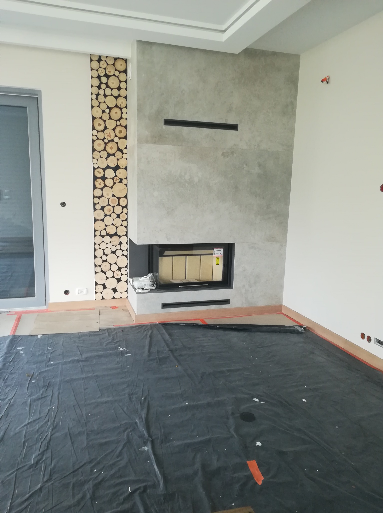 Wnętrze w trakcie prac wykończeniowych z nowoczesnym kominkiem obłożonym szarym kamieniem i dekoracyjnym panelem z przekrojów drewna na ścianie, podłoga zabezpieczona folią malarską.