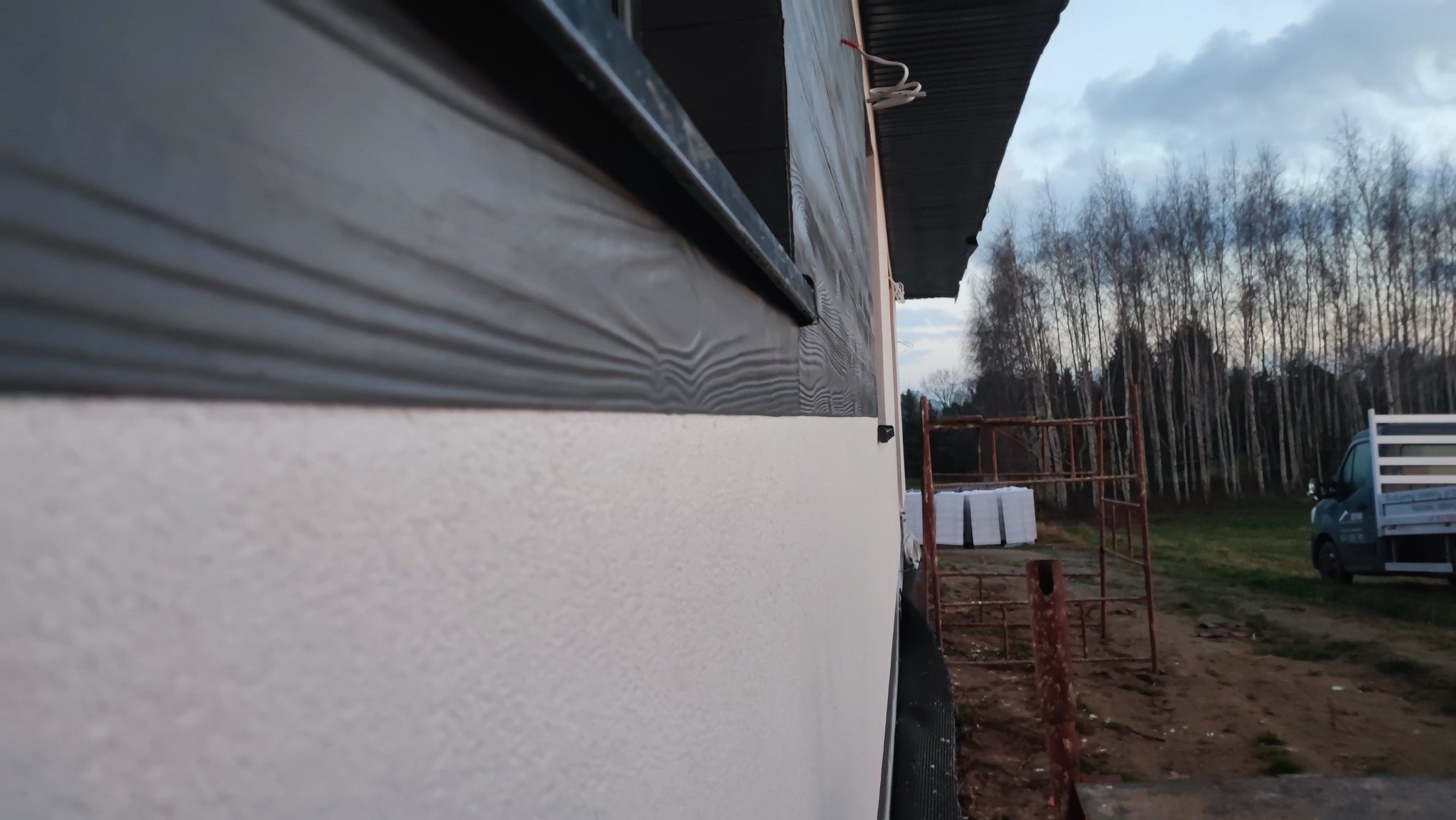 Fragment elewacji budynku z szarą okładziną imitującą drewno i białą, chropowatą ścianą, widok z boku, w tle drzewa bez liści i dostawczy samochód.