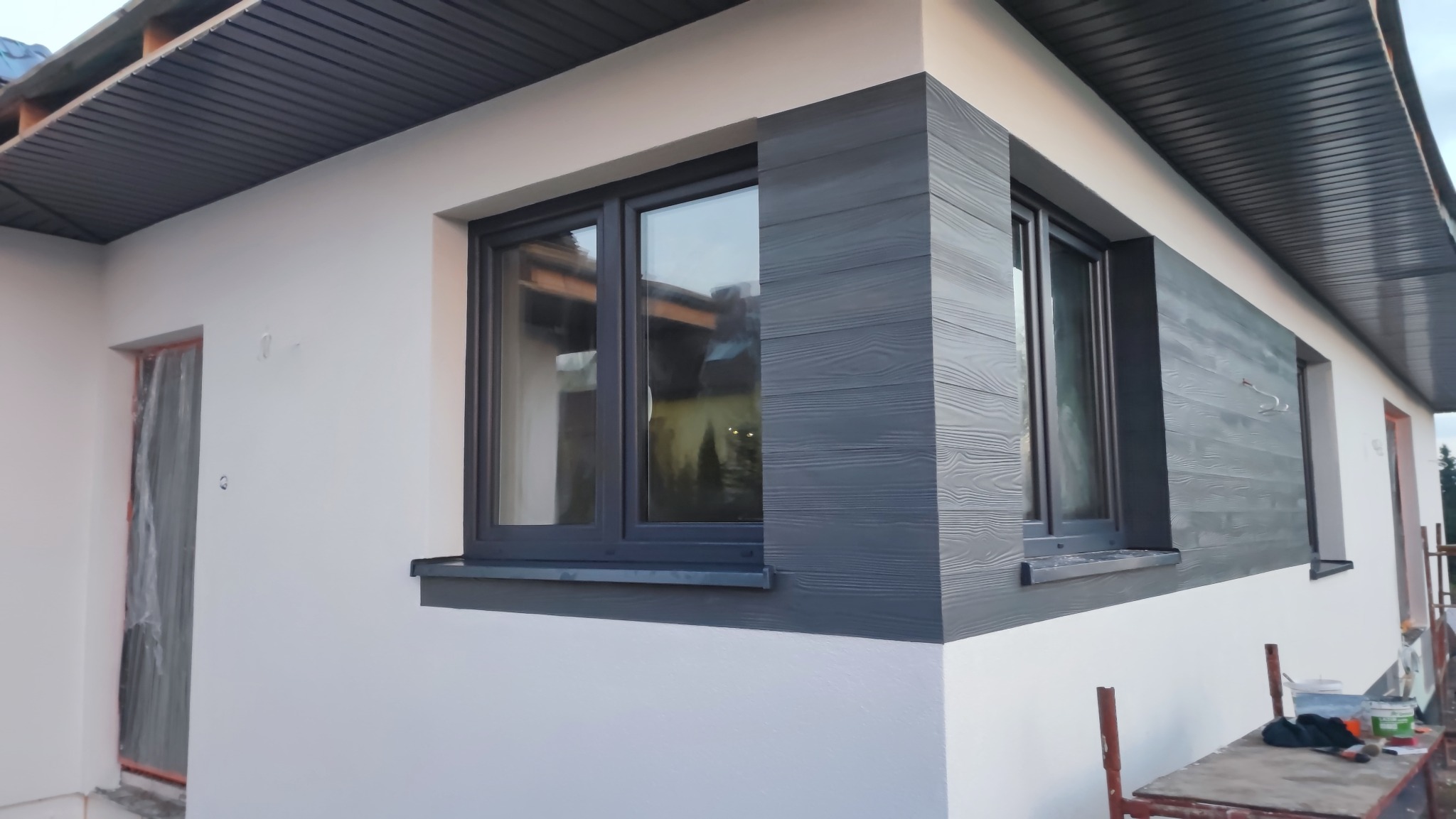 Wykończona elewacja domu z szarymi panelami imitującymi drewno wokół okien i białym tynkiem, widoczne okna z szarymi ramami, na pierwszym planie rusztowanie.