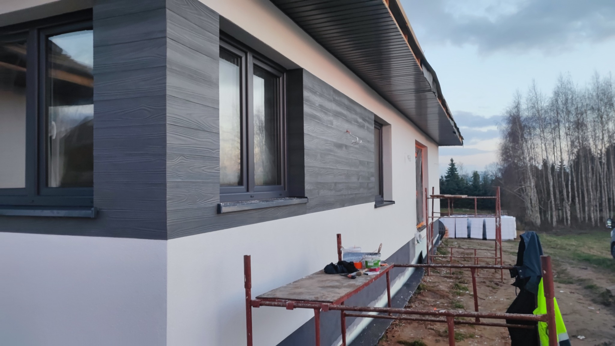 Elewacja domu jednorodzinnego z szarą okładziną imitującą drewno, widoczne rusztowanie i puszki z farbą, drzewa w tle.