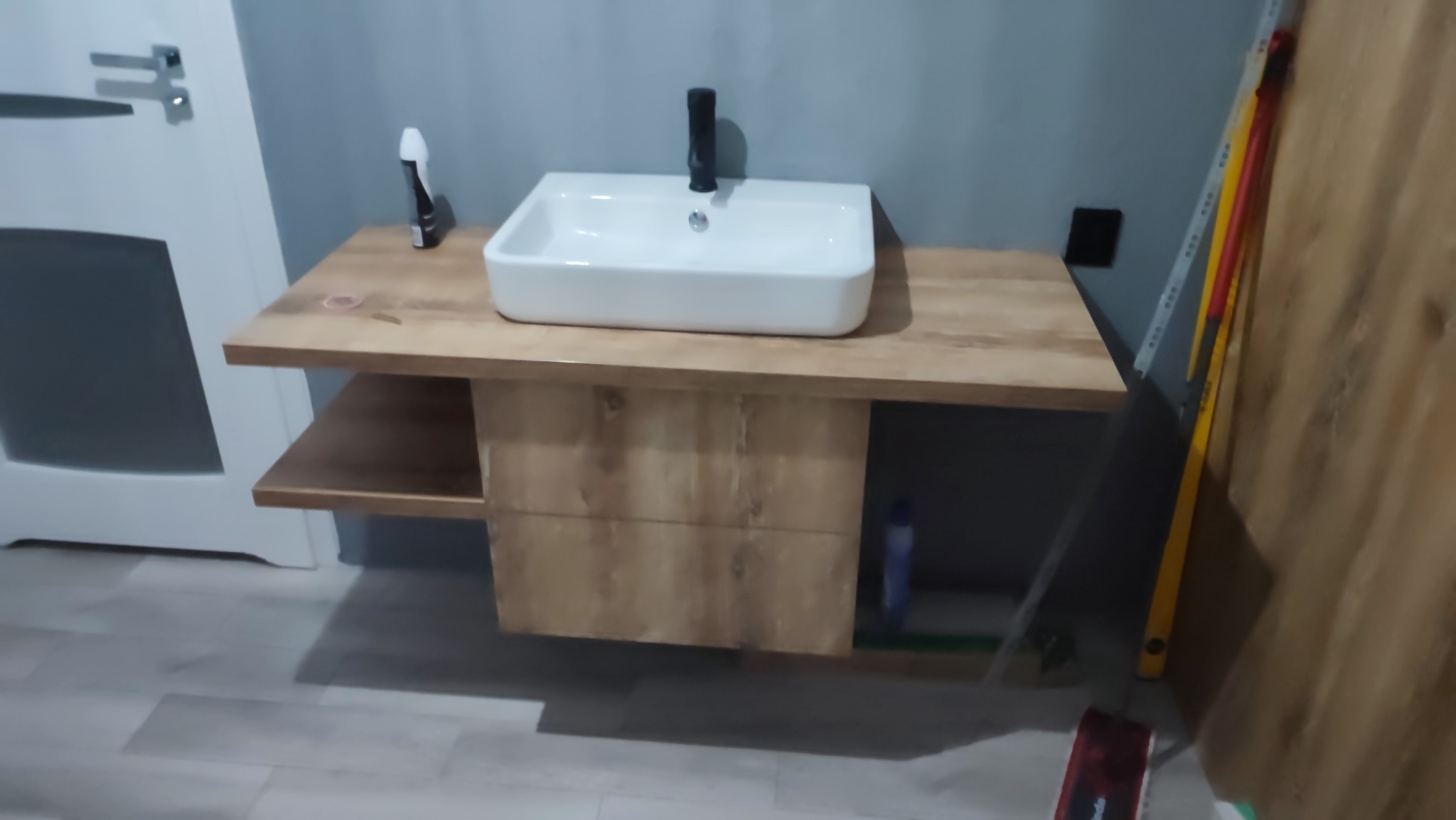 Nowoczesna, prostokątna umywalka nablatowa z czarną armaturą, zamontowana na drewnianej szafce z półką w łazience z szarą ścianą, widoczne narzędzia przy ścianie.