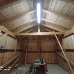 Wnętrze drewnianej konstrukcji dachu z widocznym oświetleniem LED, narzędziami na stole i fragmentem drabiny po prawej stronie.