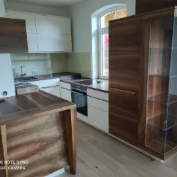 Nowoczesna kuchnia z drewnianymi i białymi szafkami, widok na okno z jesiennymi drzewami, blat z płytą indukcyjną i niezamontowane gniazdko elektryczne na ścianie.