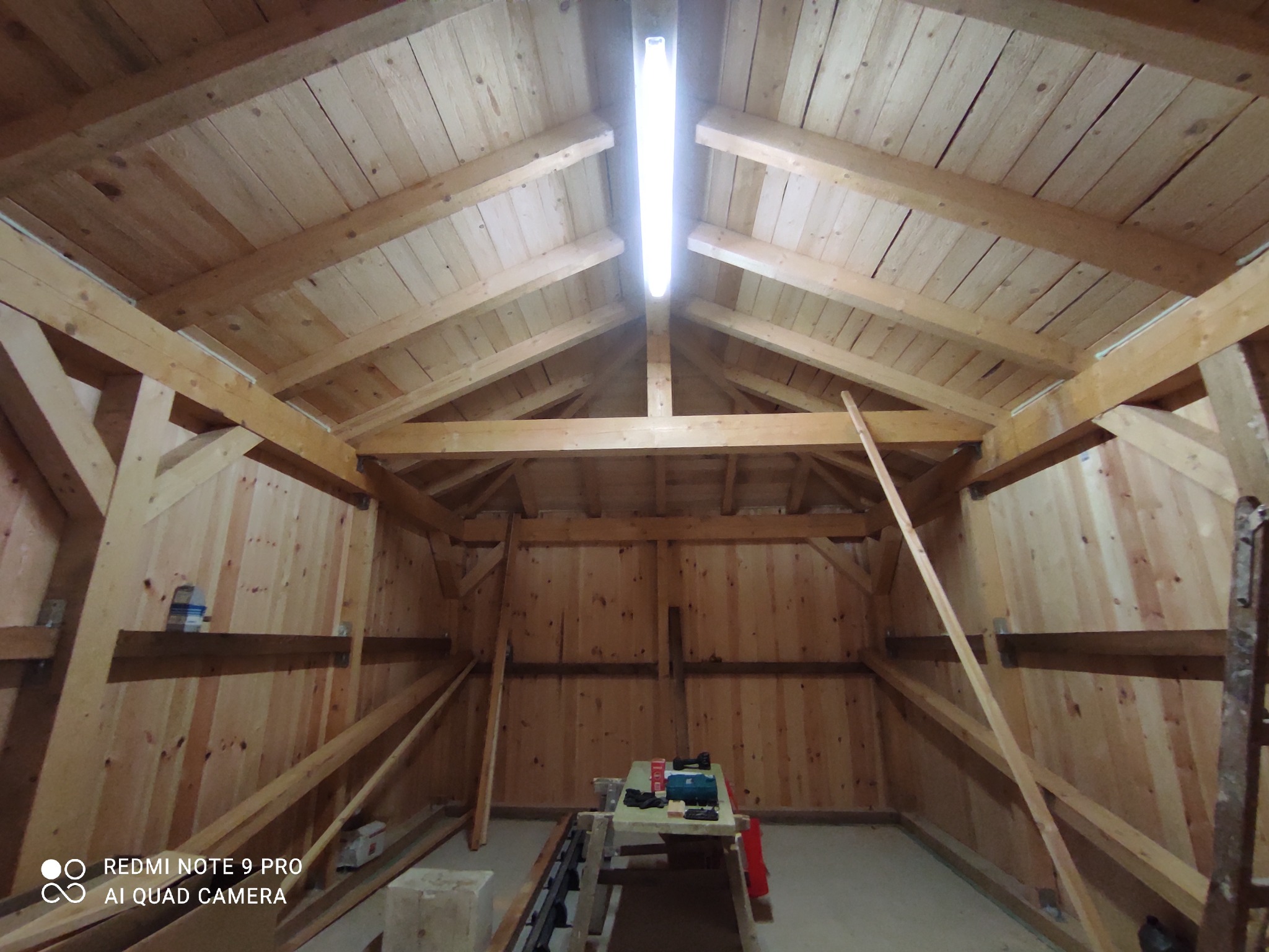 Wnętrze drewnianej konstrukcji dachu z widocznym oświetleniem LED, narzędziami na stole i fragmentem drabiny po prawej stronie.