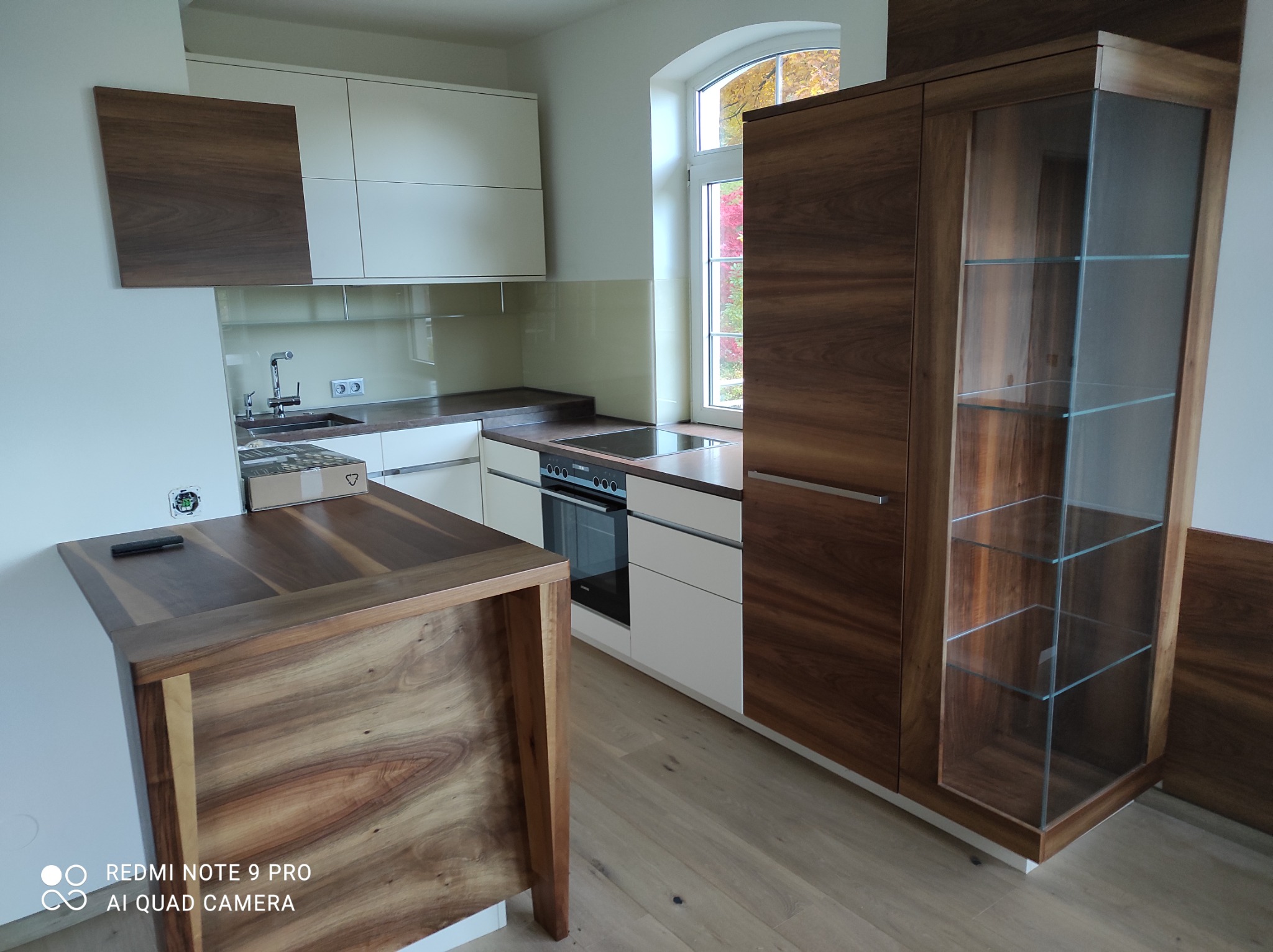 Nowoczesna kuchnia z drewnianymi i białymi szafkami, widok na okno z jesiennymi drzewami, blat z płytą indukcyjną i niezamontowane gniazdko elektryczne na ścianie.