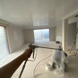 DreamWalls Maciej Zajkowski - Aplikacja gładzi szpachlowej natryskowo za pomocą agregatu malarskiego w pomieszczeniu w stanie deweloperskim; widoczny pistolet natryskowy, wiadro z gładzią Flugger i drabina.