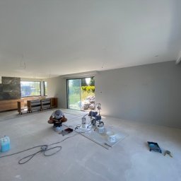 DreamWalls Maciej Zajkowski - Wnętrze domu w trakcie malowania, widoczny malarz przygotowujący farbę, agregat malarski oraz świeżo pomalowane ściany na szaro.