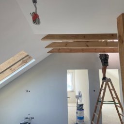 DreamWalls Maciej Zajkowski - Adaptacja poddasza: mężczyzna na drabinie pracuje przy drewnianych belkach stropowych, widoczne przygotowane ściany do dalszych prac, wentylator i wiaderka z farbą.