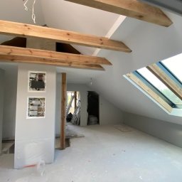 DreamWalls Maciej Zajkowski - Poddasze po remoncie z drewnianymi belkami stropowymi, oknem dachowym i częściowo zakrytą folią ścianą z otwartymi skrzynkami elektrycznymi.