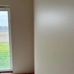 DreamWalls Maciej Zajkowski - Świeżo pomalowane ściany w odcieniach beżu, z widocznym zabezpieczeniem podłogi taśmą malarską i folią, okno z widokiem na zewnątrz.