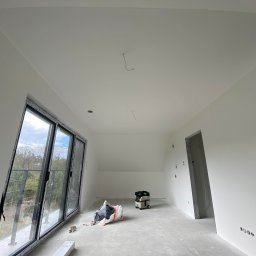 DreamWalls Maciej Zajkowski - Wnętrze nowo wykończonego pokoju z dużym oknem balkonowym, świeżo pomalowane ściany na biało, widoczny worek gruzu i odkurzacz przemysłowy, surowy betonowy węgarek drzwiowy.