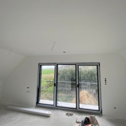 DreamWalls Maciej Zajkowski - Poddasze z oknem balkonowym i świeżo pomalowanymi na biało ścianami, widoczne puszki elektryczne bez osprzętu, surowa wylewka podłogowa i materiały budowlane.