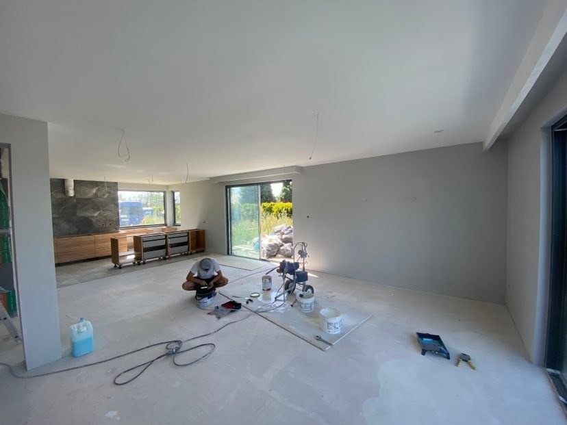 Wnętrze domu w trakcie malowania, widoczny malarz przygotowujący farbę, agregat malarski oraz świeżo pomalowane ściany na szaro.