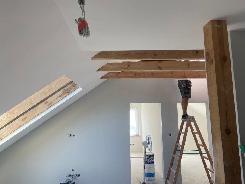 Adaptacja poddasza: mężczyzna na drabinie pracuje przy drewnianych belkach stropowych, widoczne przygotowane ściany do dalszych prac, wentylator i wiaderka z farbą.