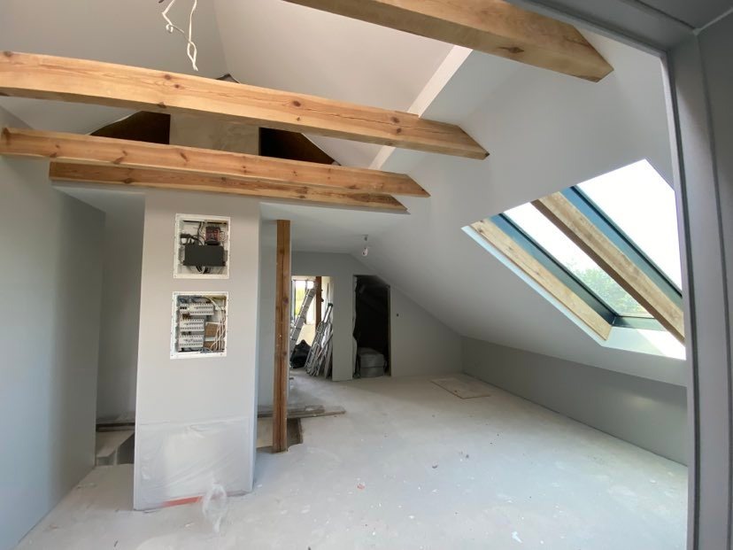 Poddasze po remoncie z drewnianymi belkami stropowymi, oknem dachowym i częściowo zakrytą folią ścianą z otwartymi skrzynkami elektrycznymi.