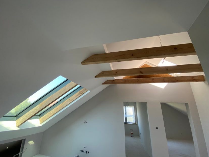 Poddasze w Warszawie po ociepleniu, widoczne drewniane belki i okno dachowe, ściany przygotowane do malowania.