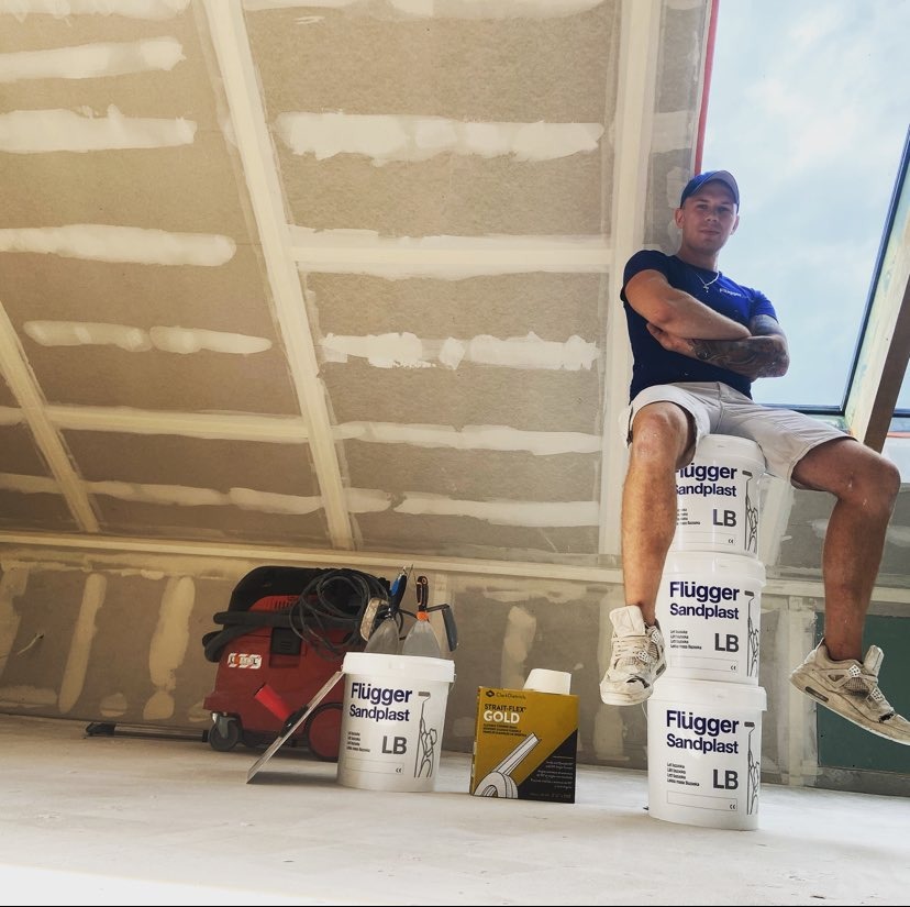 Młody mężczyzna siedzi na wiadrach z masą szpachlową Flugger Sandplast w pomieszczeniu z płytami kartonowo-gipsowymi na poddaszu, widoczny odkurzacz przemysłowy i puszka Strait Flex Gold.