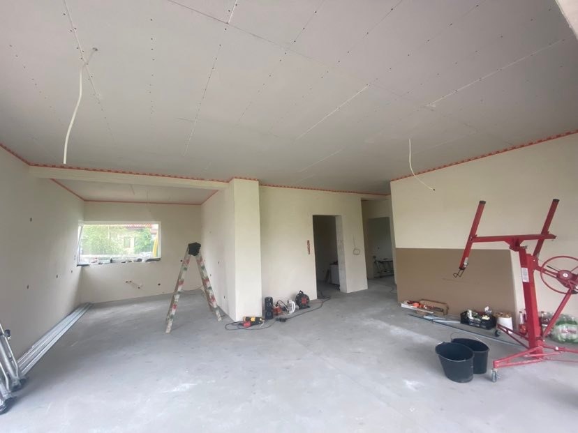 Wnętrze w trakcie montażu płyt kartonowo-gipsowych na suficie, widoczna drabina, podnośnik do płyt, profile i narzędzia na podłodze, okno z widokiem na zieleń.