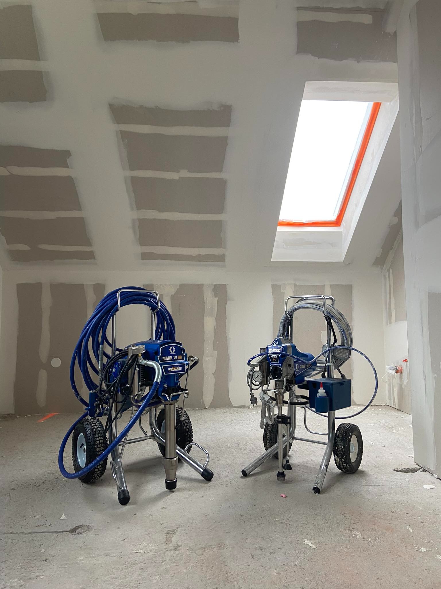 Dwa agregaty malarskie Graco z niebieskimi wężami w surowym wnętrzu z płytami kartonowo-gipsowymi i oknem dachowym, przygotowane do malowania.