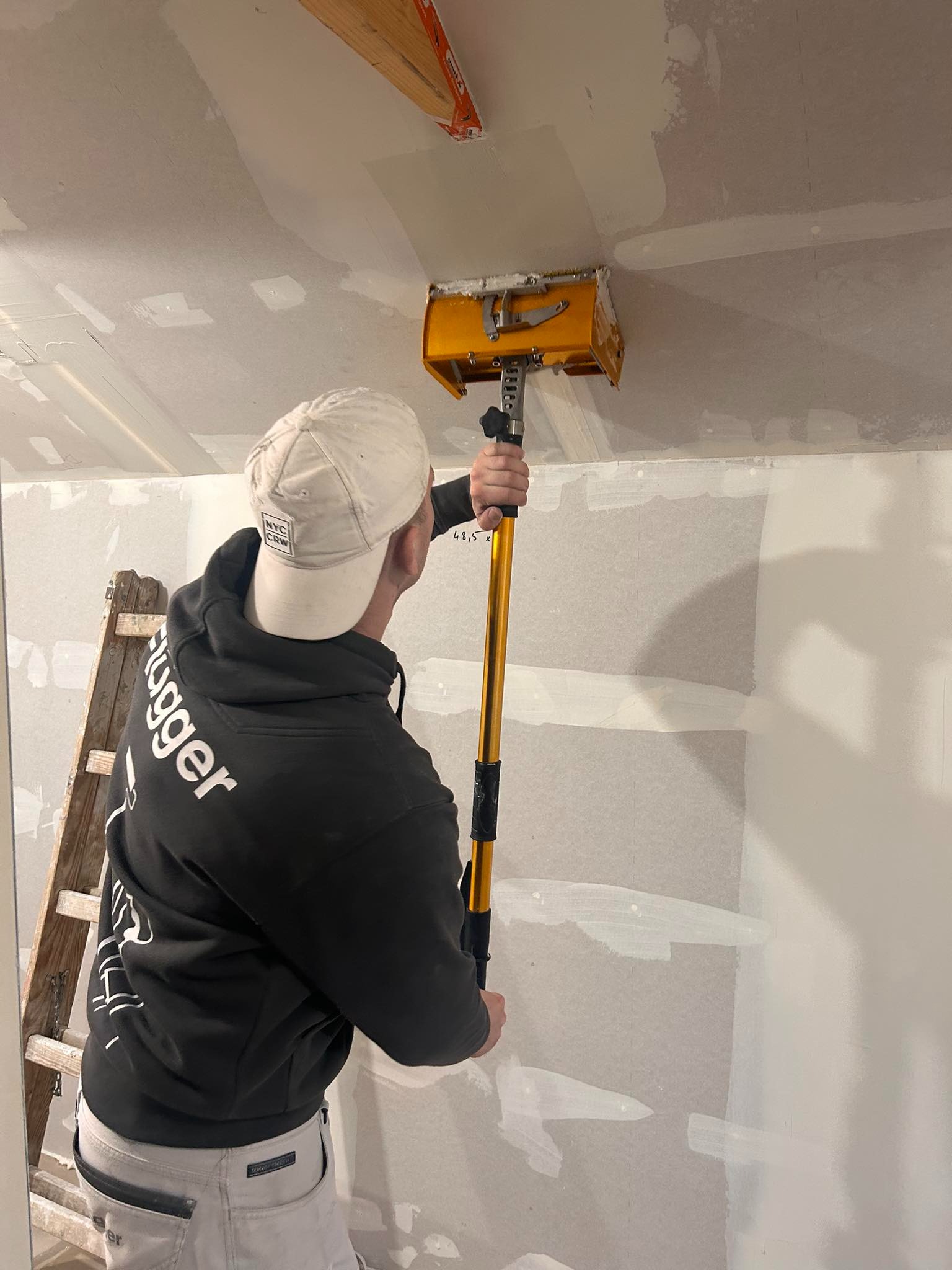 Pracownik w bluzie z kapturem, używający żółtej pacy teleskopowej do nakładania gładzi na łączenia płyt kartonowo-gipsowych na suficie, widoczna drabina malarska.