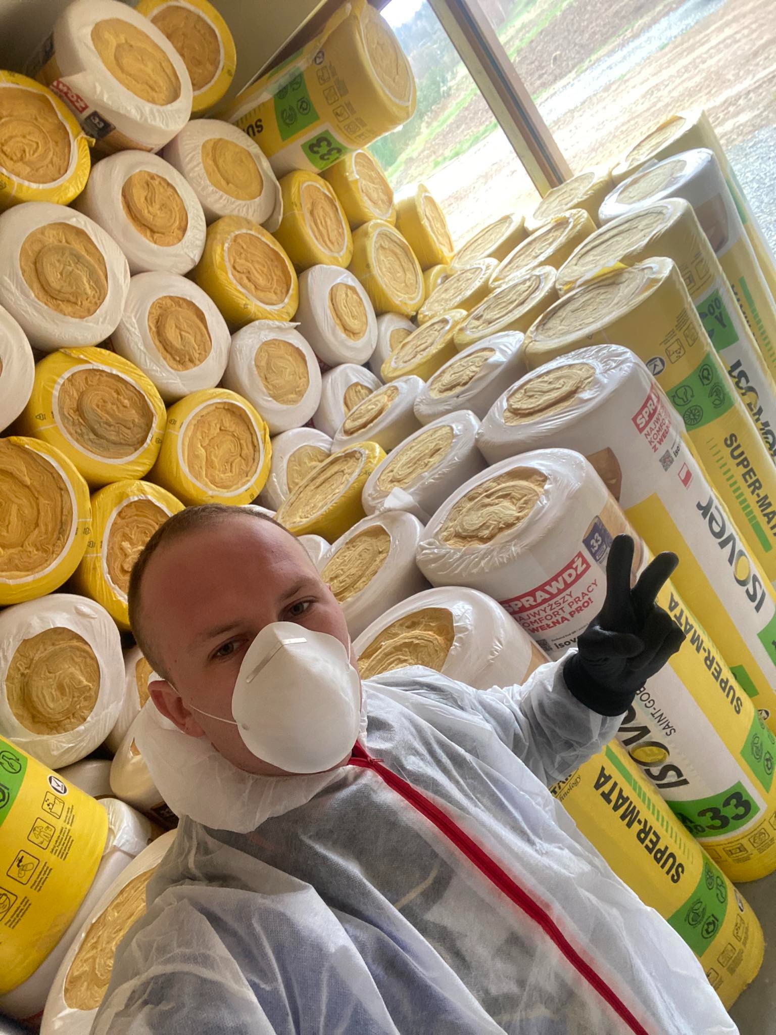 Pracownik w kombinezonie ochronnym i masce FFP2 pozuje na tle stosu wełny mineralnej Isover Super-Mata 33 w rolkach i paczkach, gestykulując dłonią w czarnej rękawicy.