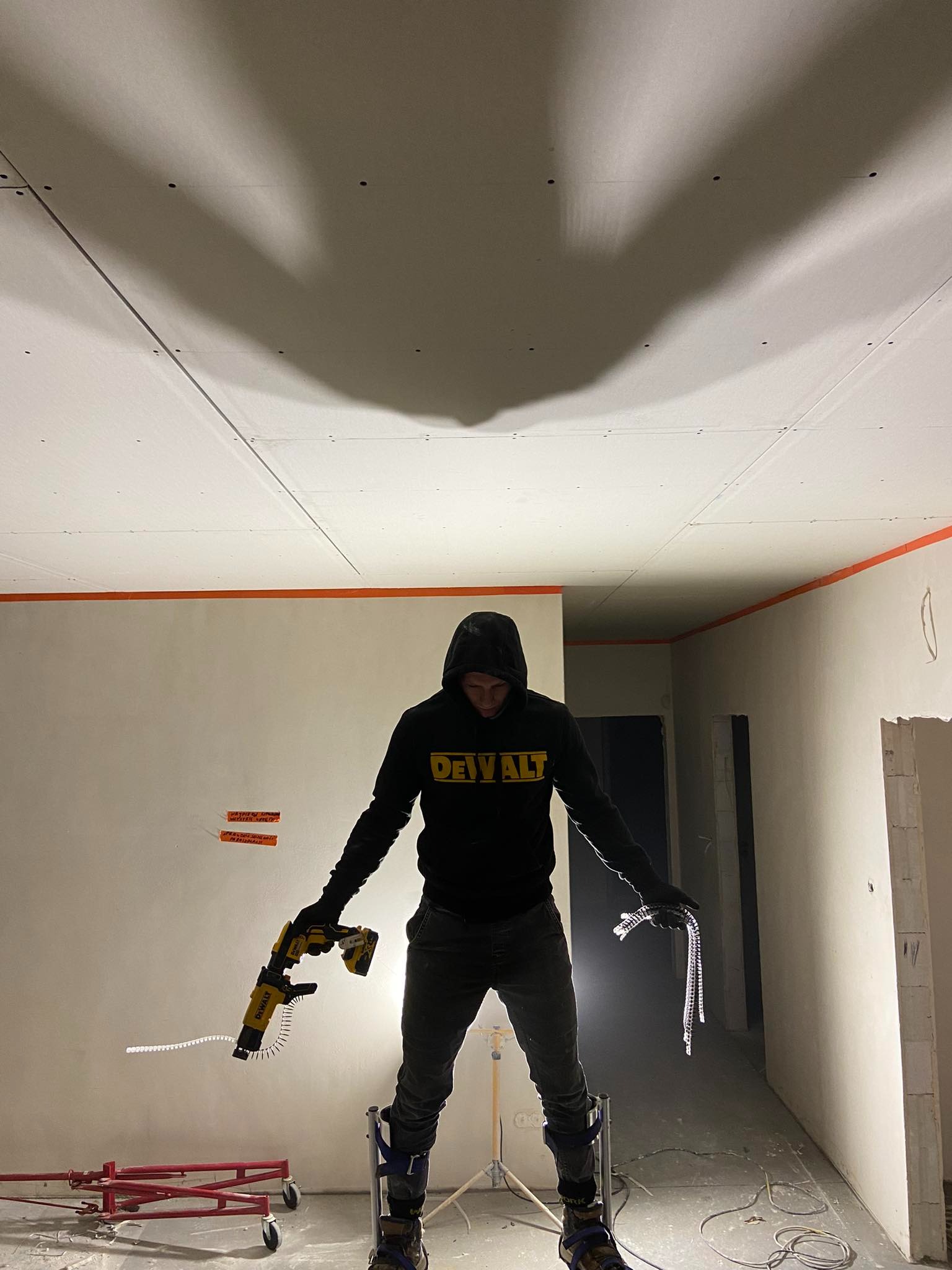 Instalator w bluzie z kapturem, na szczudłach, montuje płytę gipsowo-kartonową do sufitu za pomocą wkrętarki automatycznej DeWalt; widoczne ślady po wkrętach, pomarańczowa taśma na ścianach...