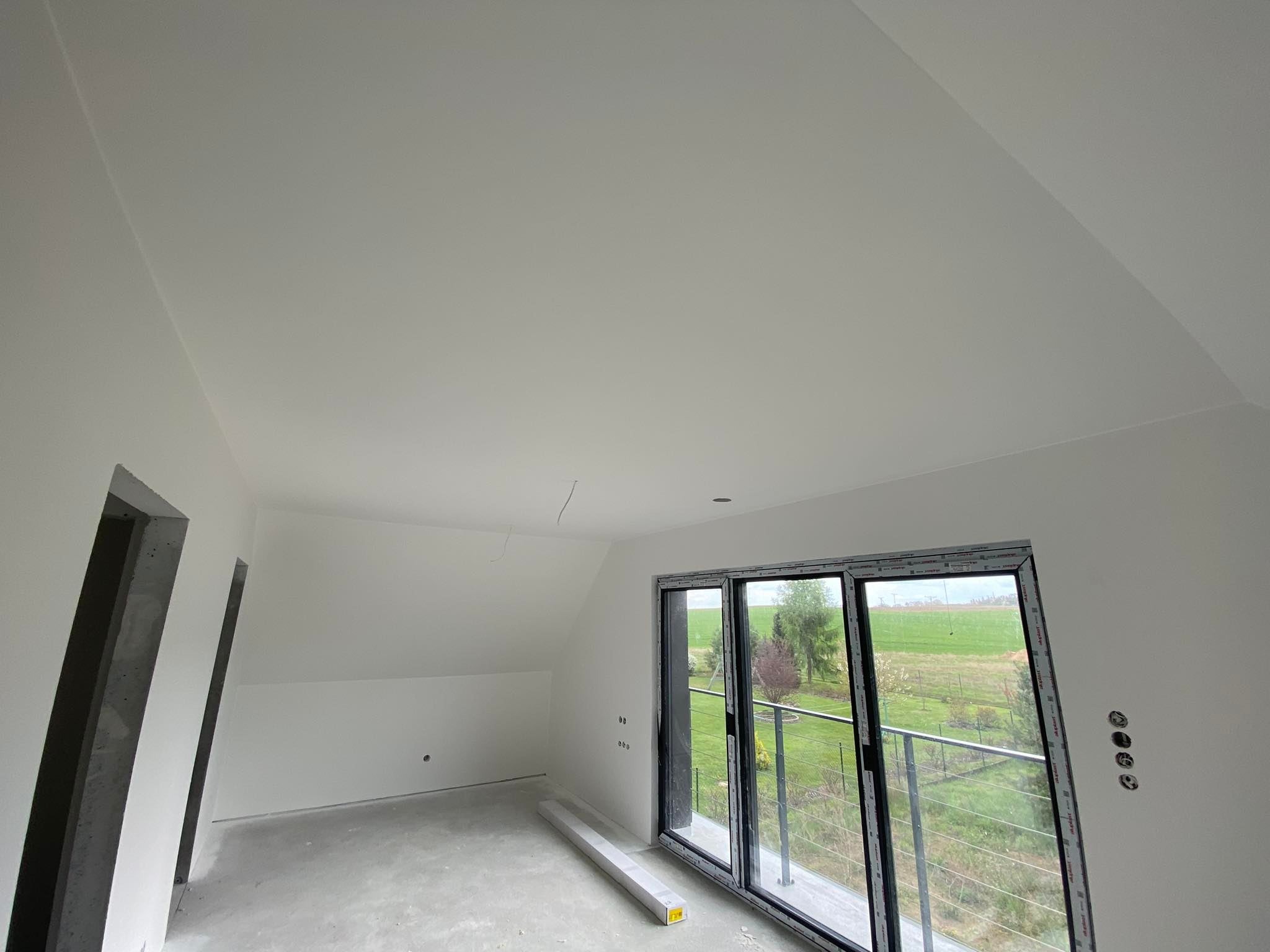 Poddasze po szpachlowaniu i malowaniu na biało, widoczne ślady po gniazdkach elektrycznych, okno balkonowe z widokiem na zieleń, surowy beton w otworach drzwiowych.