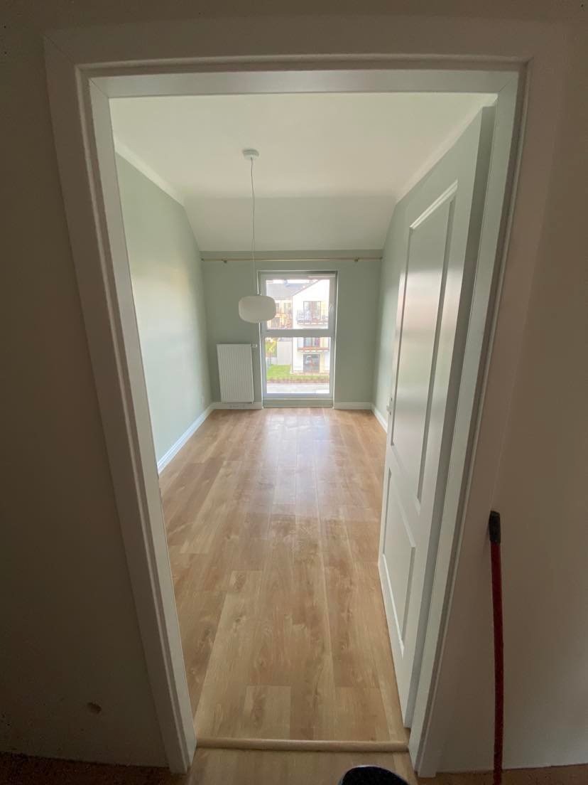 Jasne wnętrze pokoju po remoncie z drewnianą podłogą, świeżo pomalowanymi ścianami w odcieniach szarości i białymi listwami przypodłogowymi, widok przez otwarte drzwi.