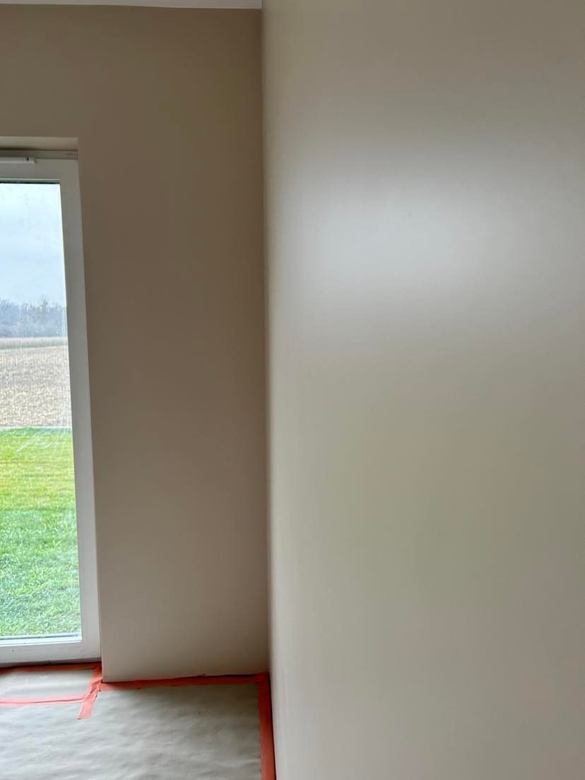 Świeżo pomalowane ściany w odcieniach beżu, z widocznym zabezpieczeniem podłogi taśmą malarską i folią, okno z widokiem na zewnątrz.
