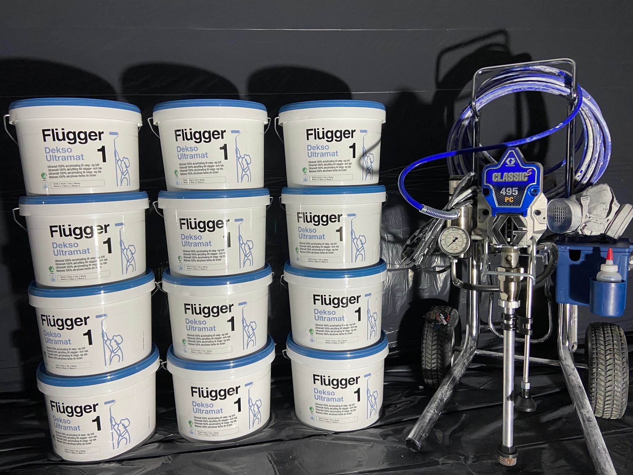 Stos puszek z białą farbą Flugger Dekso Ultramat 1, obok profesjonalny agregat malarski Classic 495 PC z niebieskim wężem, gotowy do użycia, na czarnym tle.