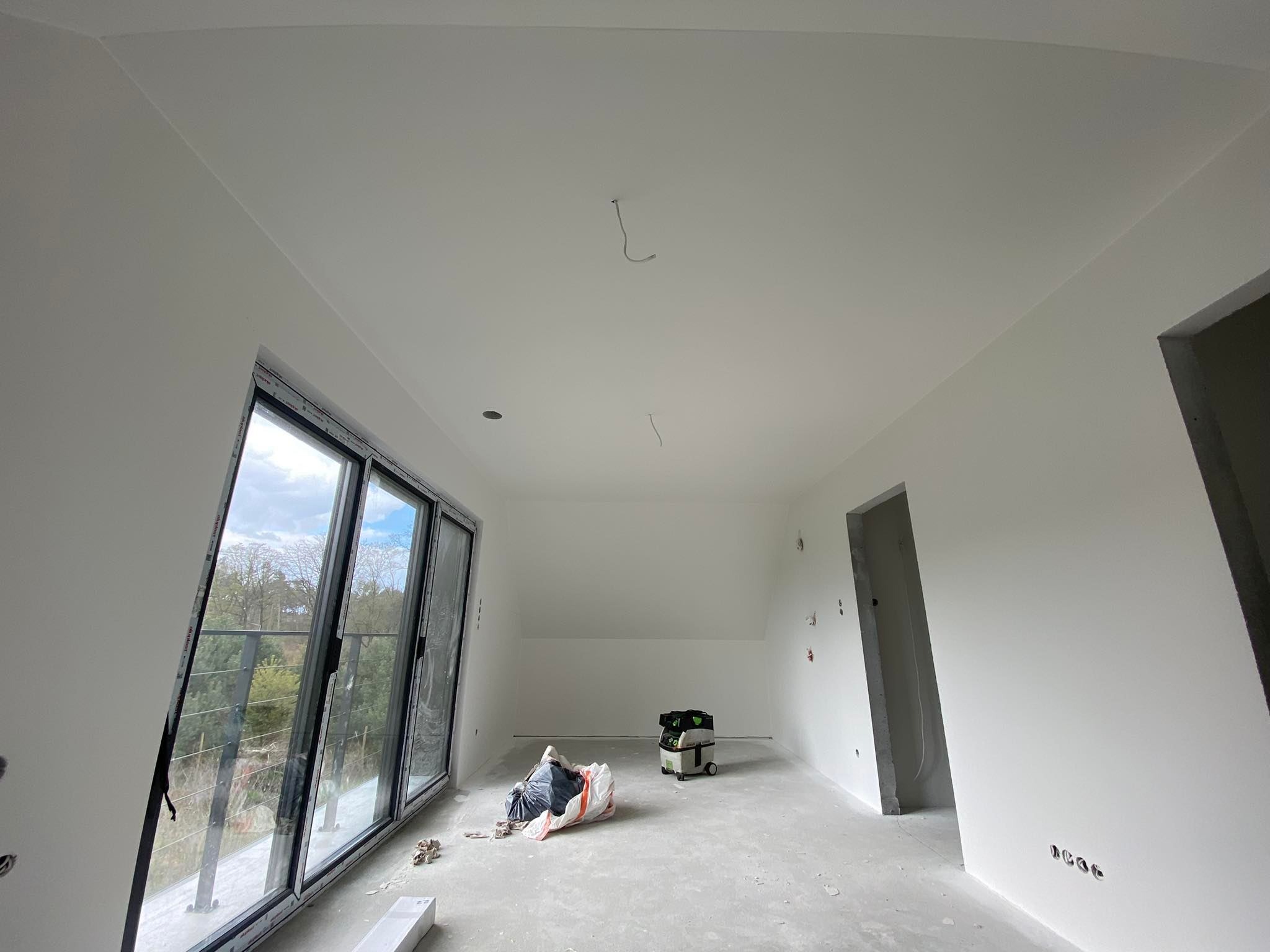 Wnętrze nowo wykończonego pokoju z dużym oknem balkonowym, świeżo pomalowane ściany na biało, widoczny worek gruzu i odkurzacz przemysłowy, surowy betonowy węgarek drzwiowy.