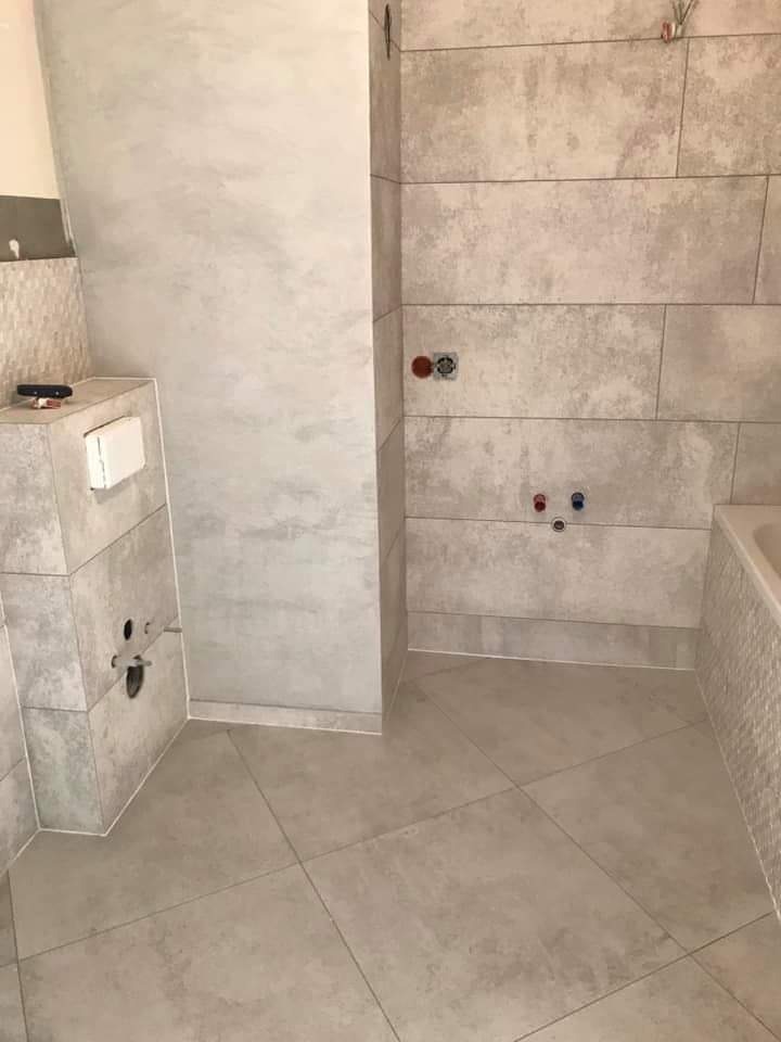 Wnętrze łazienki w trakcie remontu, widoczne ułożone płytki imitujące kamień na ścianach i podłodze, surowa ściana przygotowana pod malowanie, wystające rury instalacji wodnej i kanalizacyjnej.